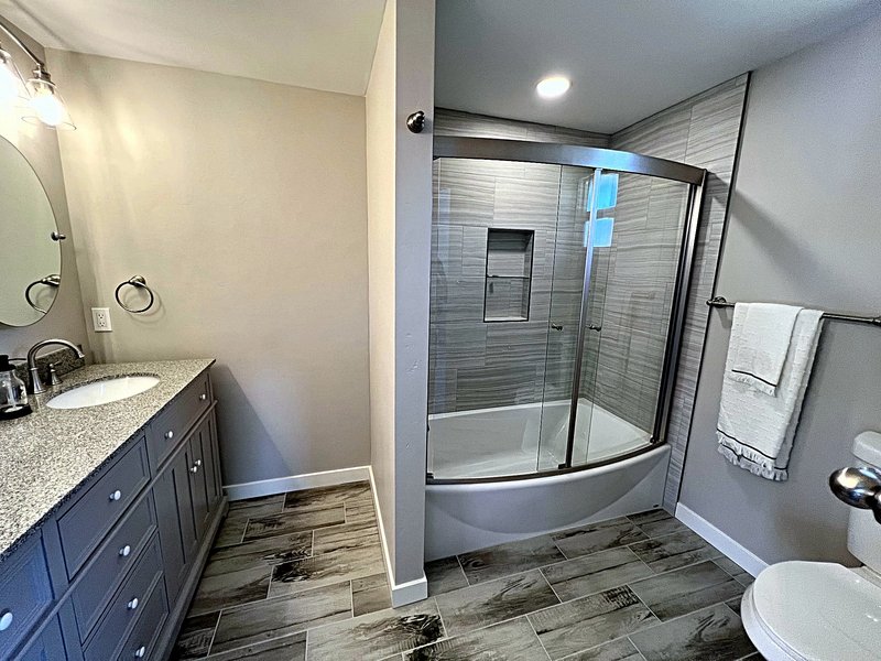 Bathroom remodel with a gray vanity and granite countertop beside a tub with a curved sliding glass door, striated tile surround, and a built-in niche
