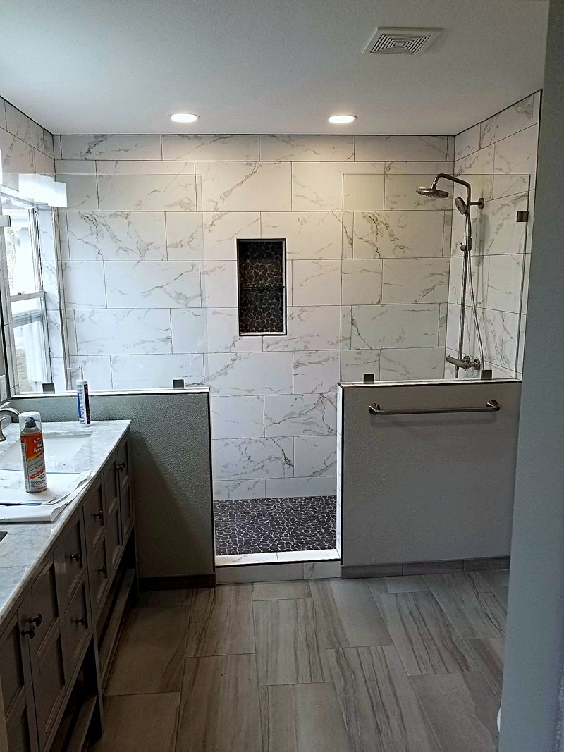Curbless walk-in shower in marble-look tile with a dark pebble floor, mosaic niche, and pony wall, next to a gray double vanity