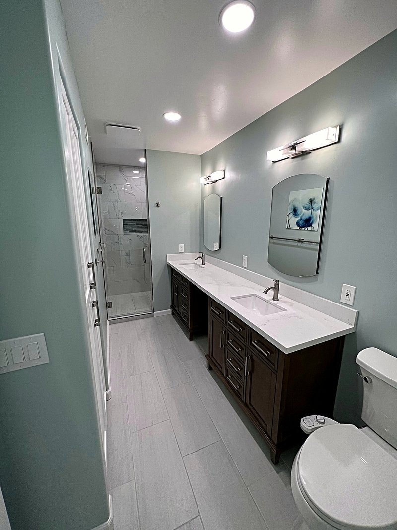 Primary bathroom we remodeled with sage-green walls, a dark wood double vanity with white quartz countertop, and a marble-look tile shower behind a glass door