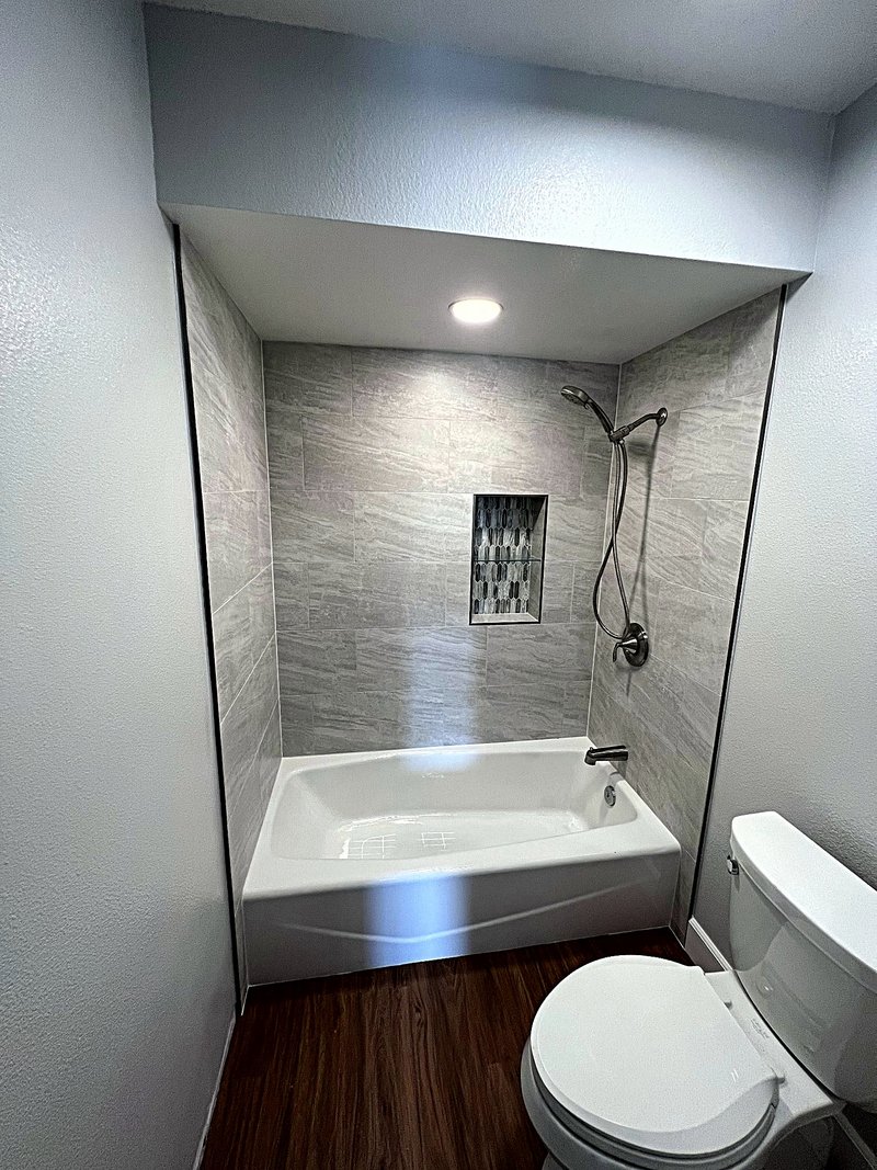 Bathroom remodel with a new alcove tub surrounded by large-format gray tile, a mosaic-tile niche with a glass shelf, handheld shower, and wood-look plank flooring