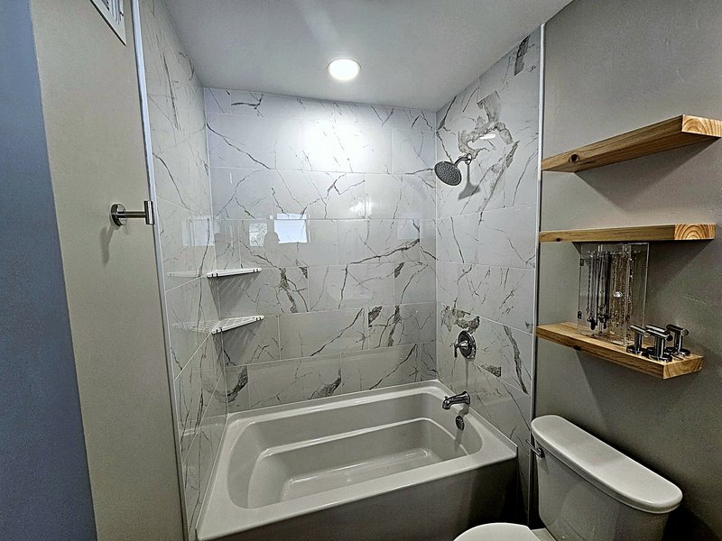 Bathroom remodel with a marble-look tile tub/shower with corner shelves, chrome fixtures, and natural wood floating shelves by the toilet