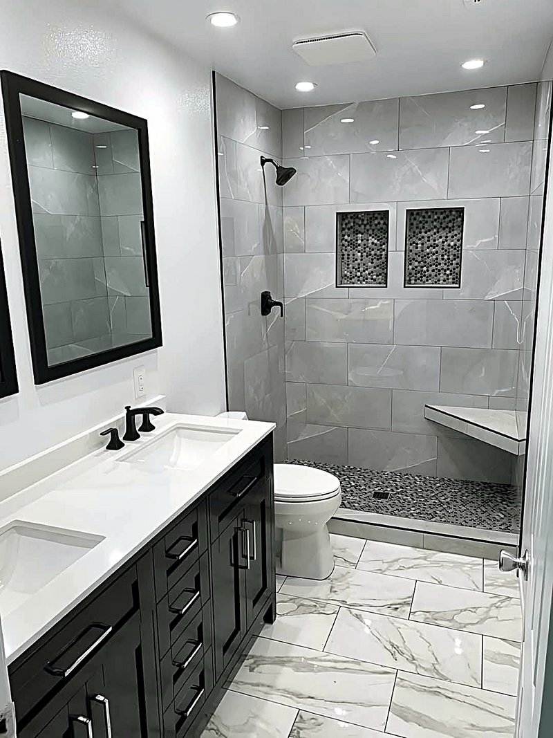 Walk-in shower in large-format gray marble-look tile with two mosaic-tiled niches, a floating corner bench, pebble shower floor, and a black double vanity