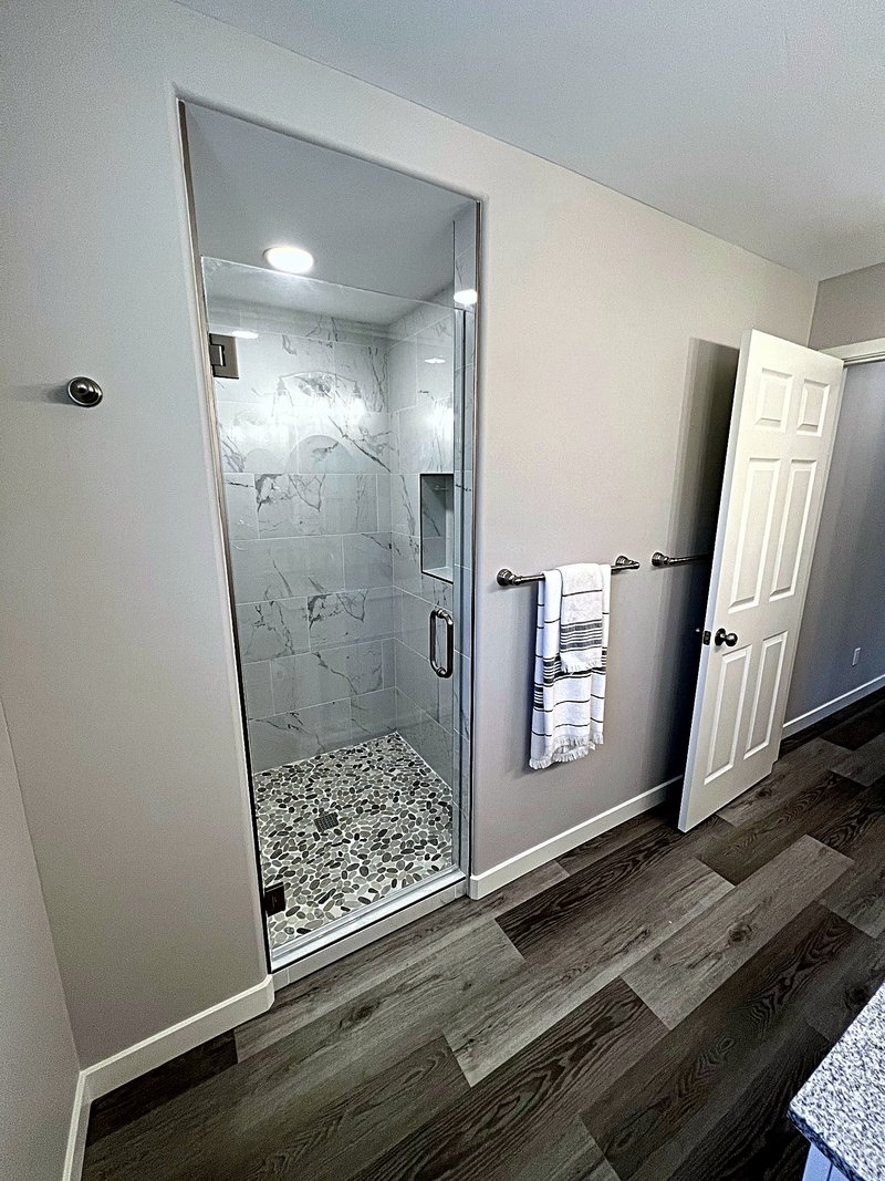 The same marble-look shower seen through its frameless glass door, with a towel bar beside it and luxury vinyl plank floors