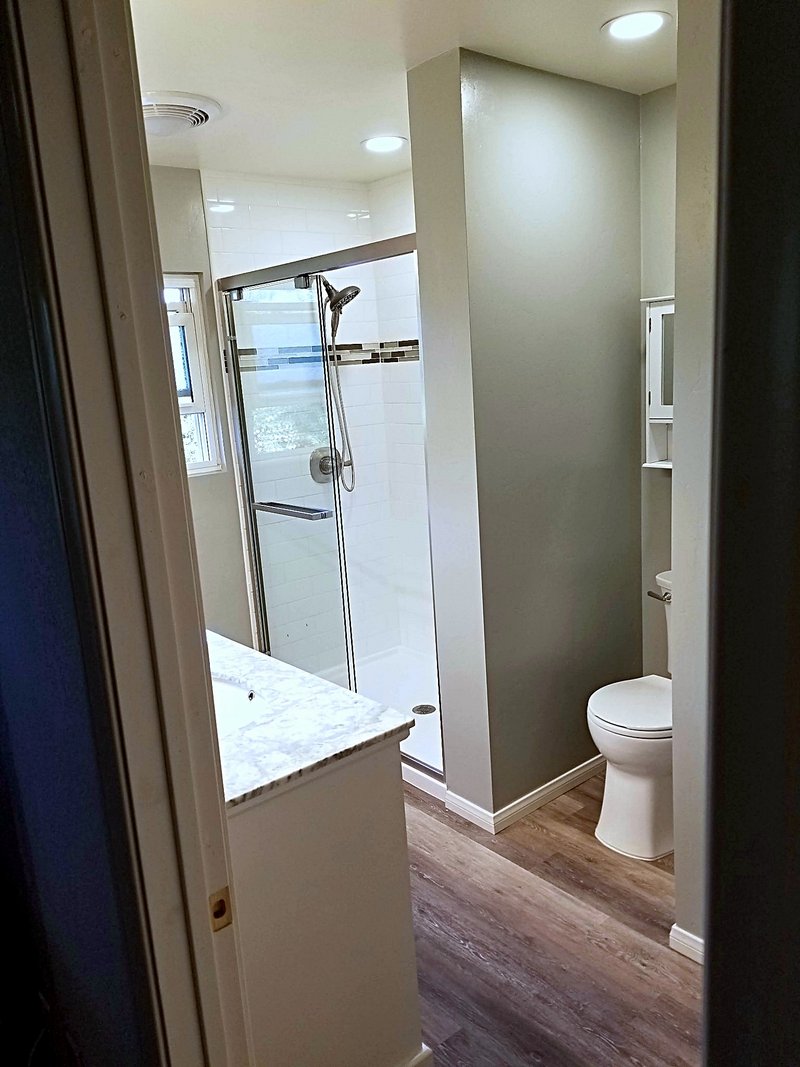 Bathroom remodel with a white subway tile shower with a mosaic accent stripe behind a sliding glass door, seen from the doorway past the vanity