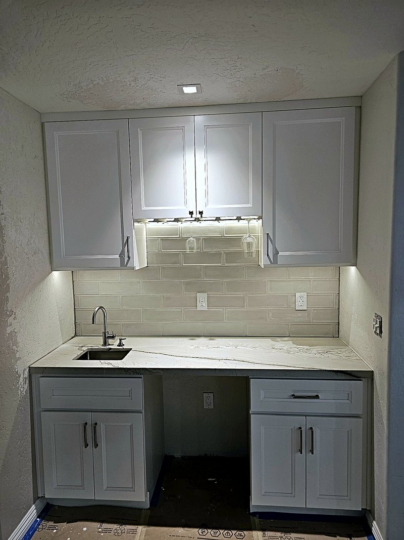 White shaker kitchenette and wet bar we built with a quartz countertop, beveled subway tile backsplash, under-cabinet lighting, wine glass rack, and a small bar sink