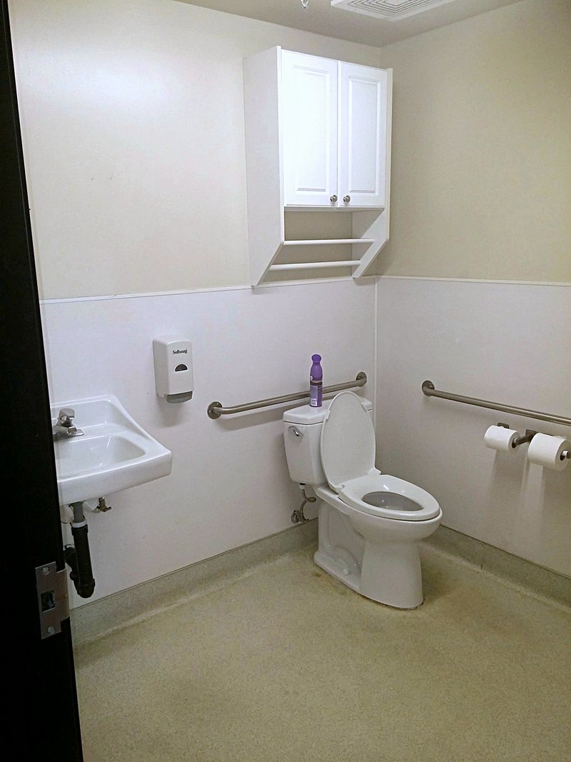 The bathroom before remodeling: plain beige walls, a basic wall-hung sink, standard toilet, and grab bars