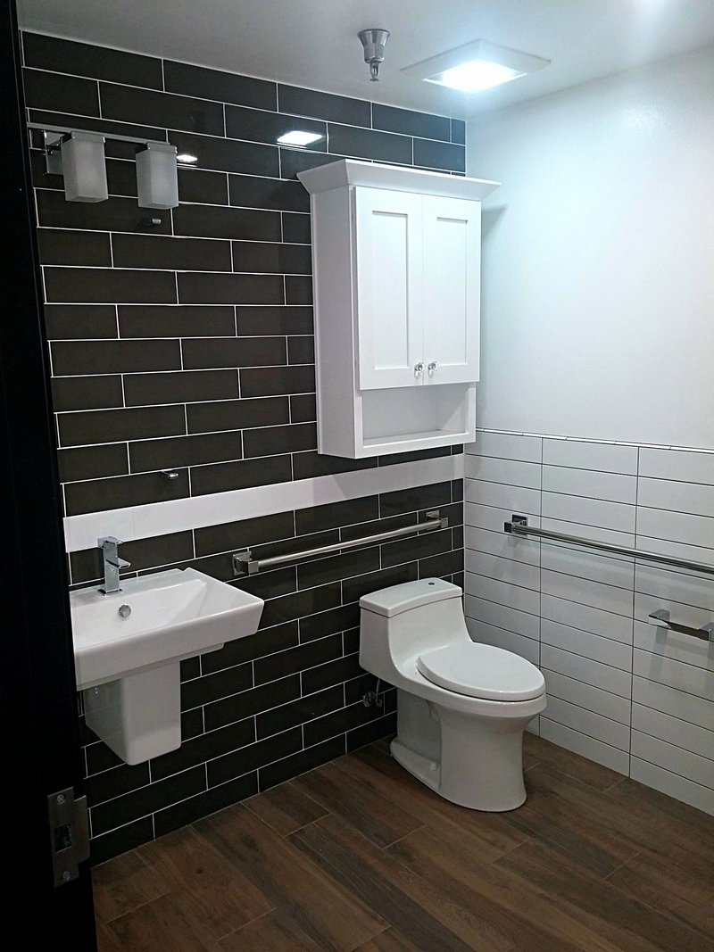 The same bathroom after remodeling: glossy black subway tile with a white accent stripe, new wall-hung sink, one-piece toilet, and wood-look plank flooring