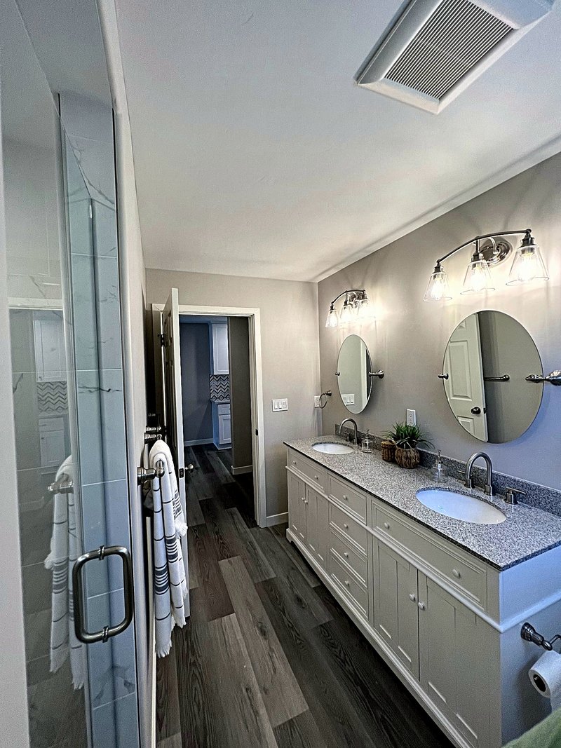 Bathroom remodel with a white double vanity, speckled granite countertop, round pivot mirrors, and a glass shower door, with the chevron-tile kitchen visible through the doorway