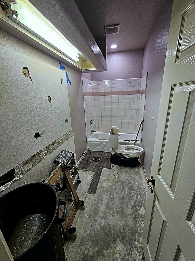 Bathroom mid-demolition: the vanity and flooring are removed and the toilet pulled, with the old tub and tile surround still in place