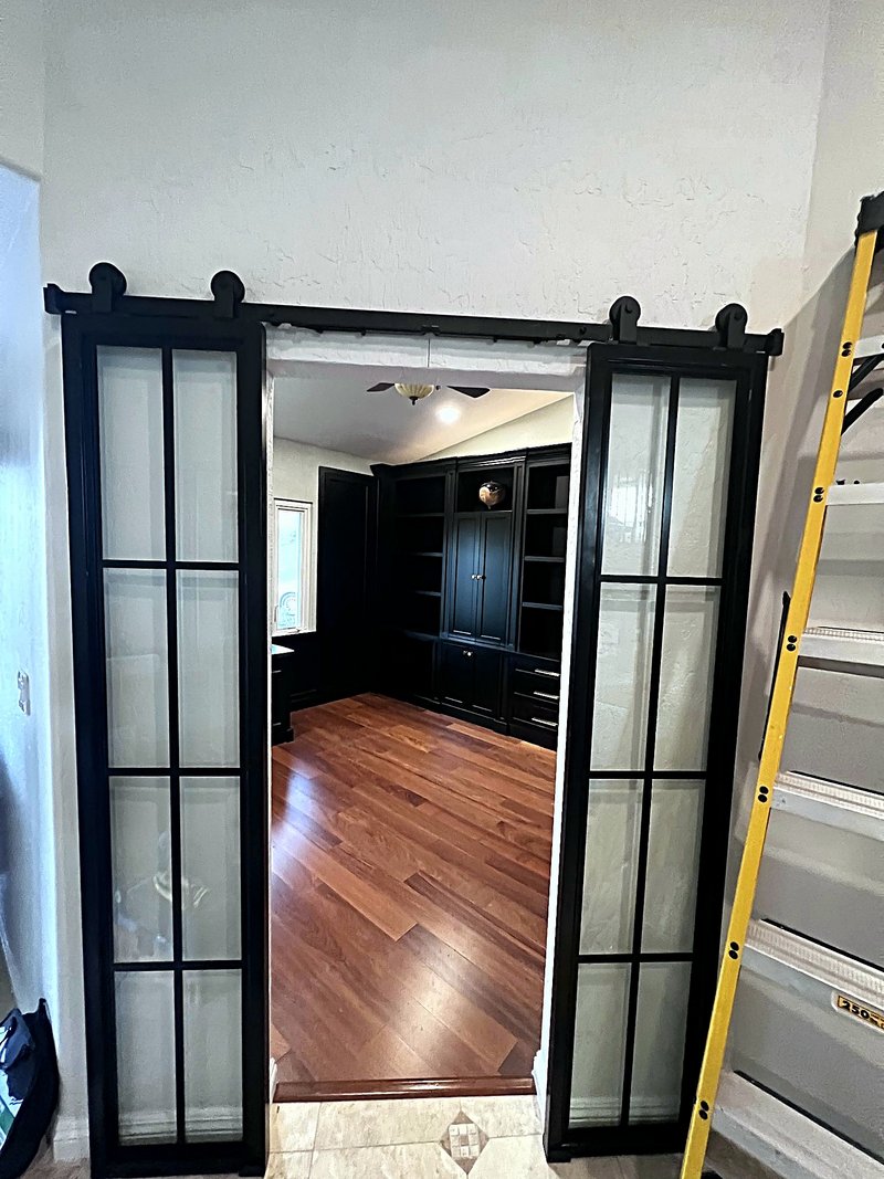 Black-framed frosted-glass sliding barn doors opening into the home office, with the black built-in bookshelves and hardwood floor visible inside