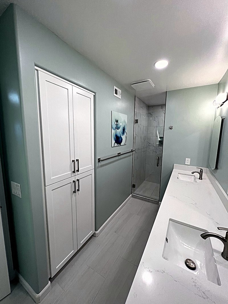 Second view of the sage-green bathroom project: a white built-in linen tower, double undermount sinks in a white quartz countertop, and the glass shower at the far end