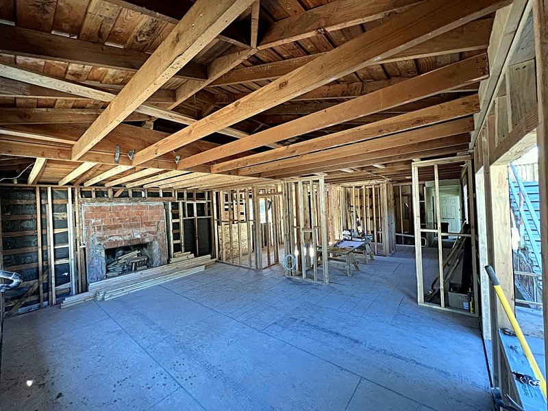 A gut renovation at the framing stage: exposed wall studs and ceiling rafters, new subfloor, and the original brick fireplace that stays in the new layout