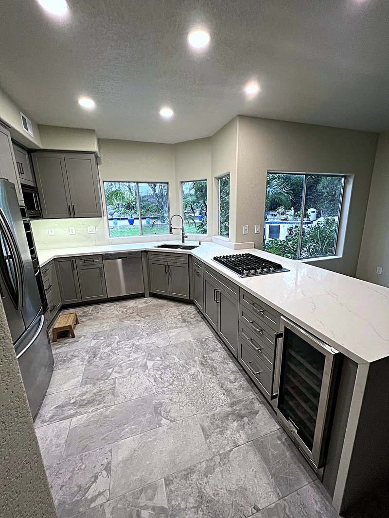 Second view of the gray shaker kitchen: peninsula with a gas cooktop, built-in wine fridge, corner sink under garden windows, and stone-look tile floors