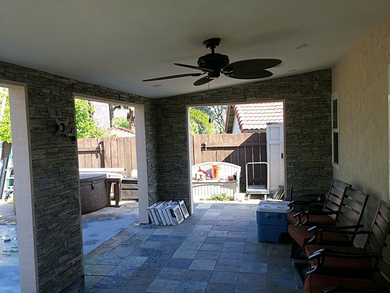 Inside the covered patio: stacked-stone walls, slate tile floor, ceiling fan, and seating, looking out to the yard