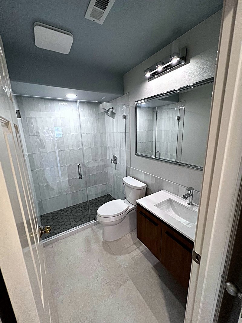 Bathroom remodel with a white tile walk-in shower behind frameless glass, charcoal hexagon shower floor, walnut vanity with a white countertop, and a wide framed mirror