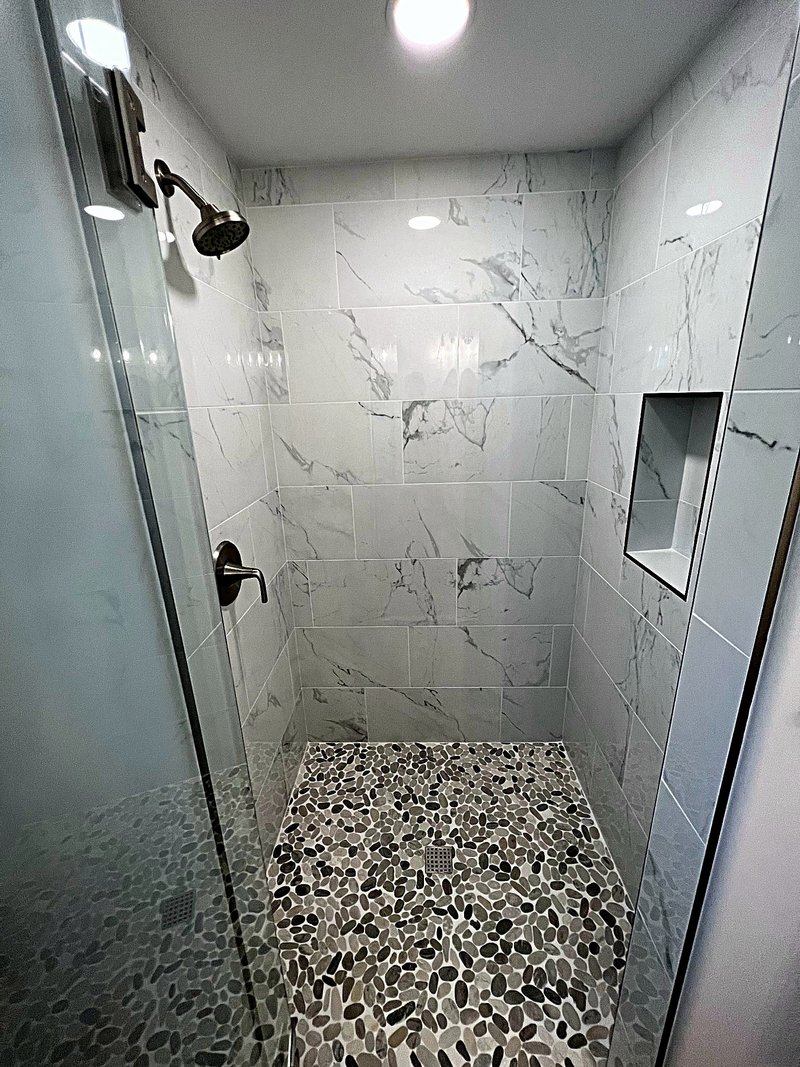 Inside view of a walk-in shower in marble-look tile with a river-rock pebble floor, recessed niche, and brushed nickel fixtures behind a glass door