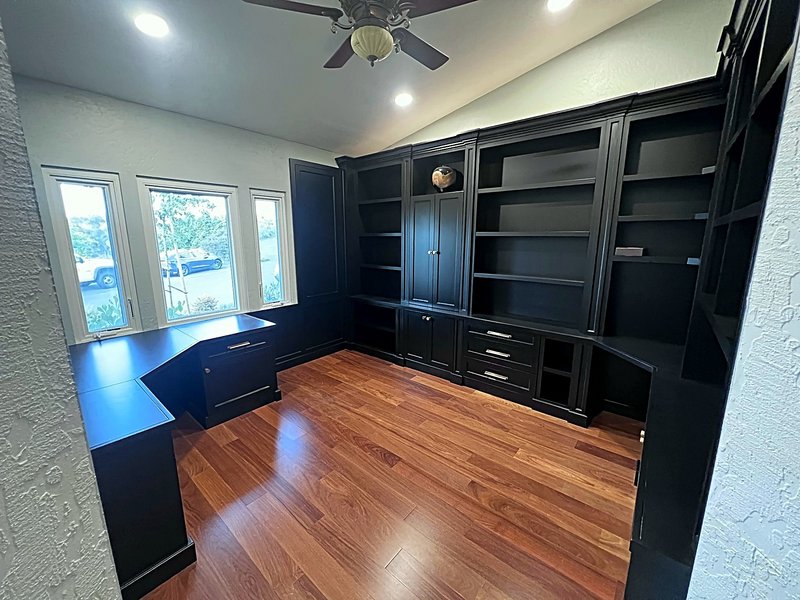 Home office with floor-to-ceiling black built-in bookshelves, cabinets and drawers, a built-in desk under three windows, and warm hardwood floors