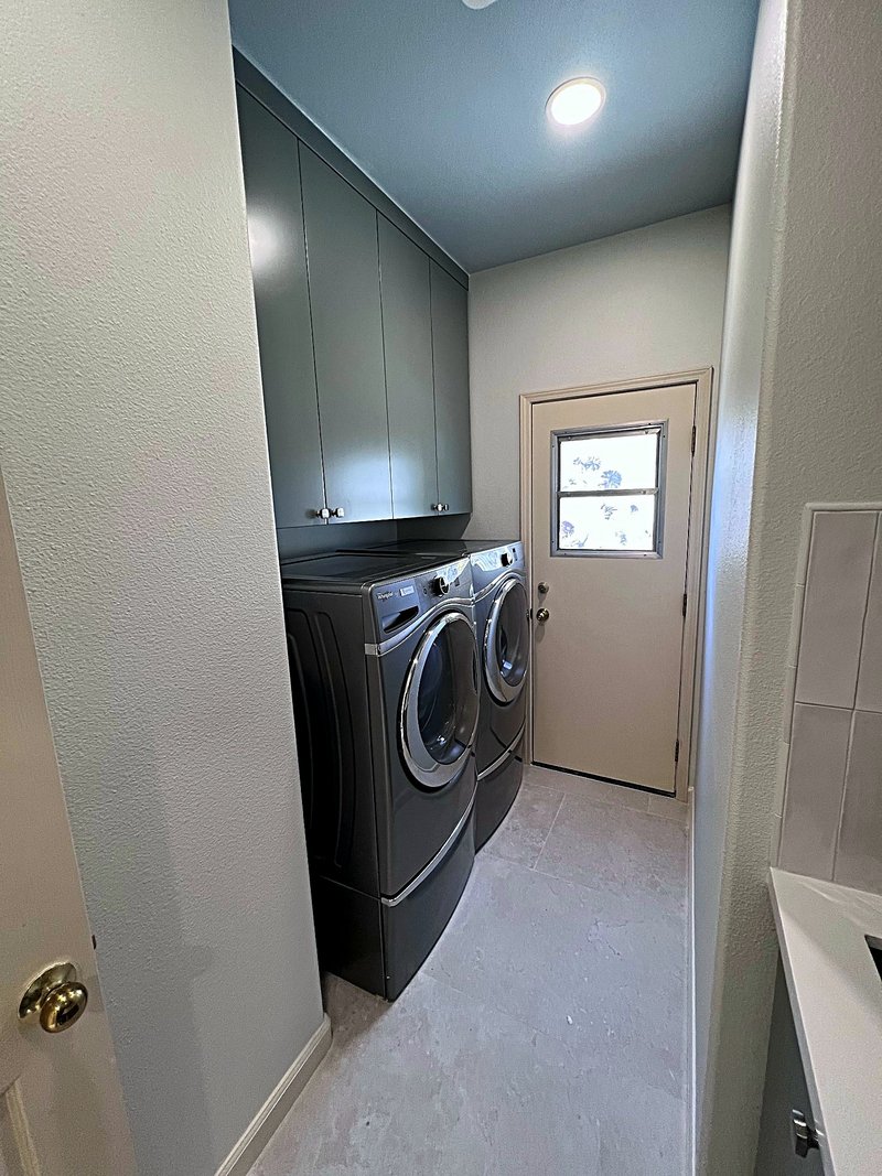 Laundry room with dark gray overhead cabinets above a front-load washer and dryer pair, large-format tile floor, and an exterior door