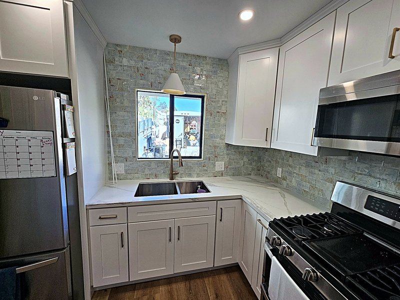 The sink wall of the same kitchen: double undermount sink with a brass faucet under the window, sea-glass subway tile, and quartz counters