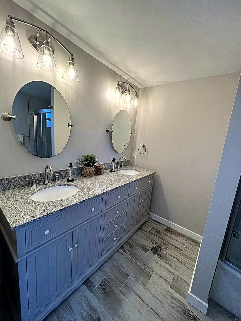 Second view of the gray bathroom: a double vanity with round pivot mirrors, bell-jar vanity lights, and wood-look tile floors, with the curved-glass shower reflected in the mirror