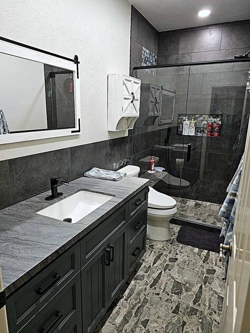 Bathroom remodel with a charcoal tile glass shower with a mosaic niche, dark vanity with stone-look countertop, and pebble-look floor tile