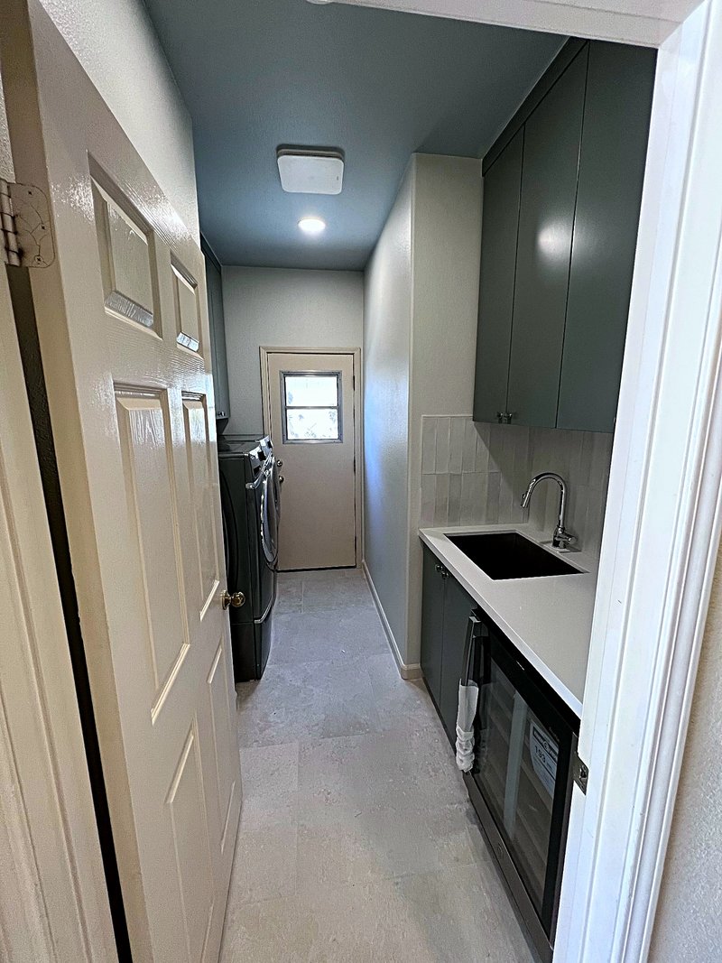 Second view of the laundry room: a black undermount utility sink in a white countertop with dark gray cabinets above and below, subway tile backsplash, and a built-in wine fridge