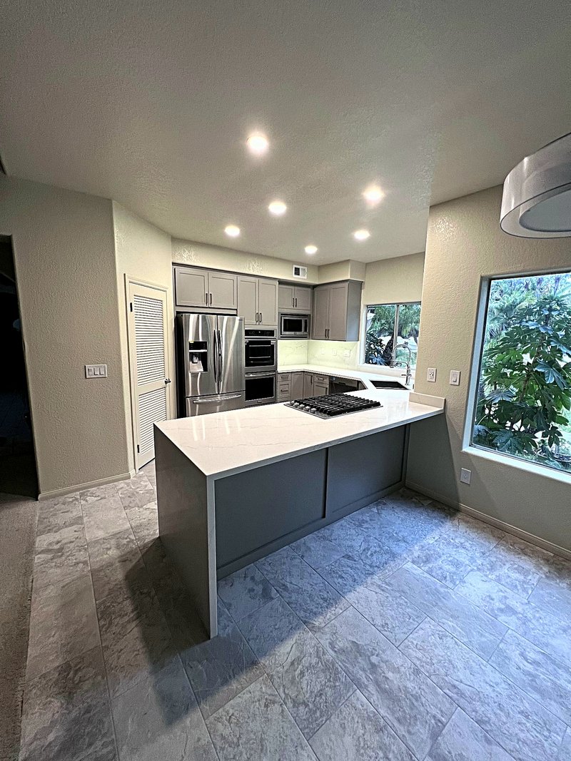 Third view of the gray shaker kitchen from the dining area: quartz waterfall island, stainless French-door refrigerator, double wall ovens, and recessed lighting