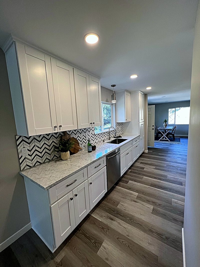 The sink wall of the same white shaker kitchen: chevron marble backsplash, long quartz countertop run, undermount sink, and tall pantry cabinets